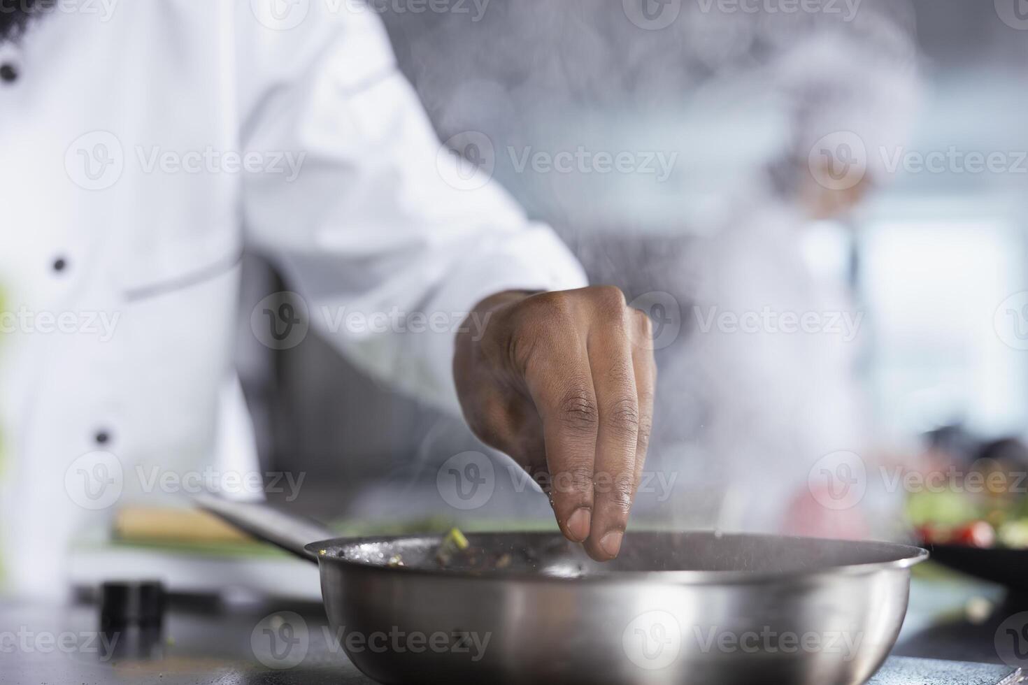 Black chef in modern kitchen adding herbs leaves to a dish on the stove, using fresh greenery and spices to elevate the flavors while cooking meal. Delicious culinary arts, gastronomy. photo