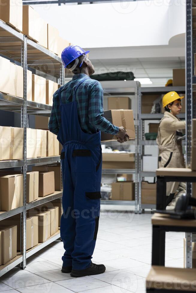 Inventory system managed by african american worker in safety gear, checking the cargo boxes on the racks. Storage units hold packages and containers for delivery, import export. photo