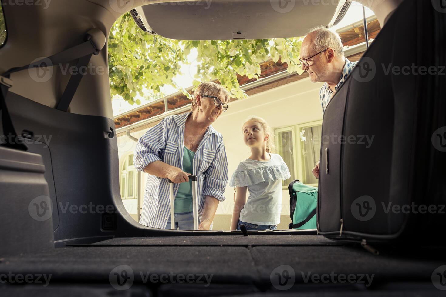 Grandparents couple taking their small niece on a summer road trip, loading the car with luggage for an exciting holiday. Senior people and little kid preparing to start the journey. photo