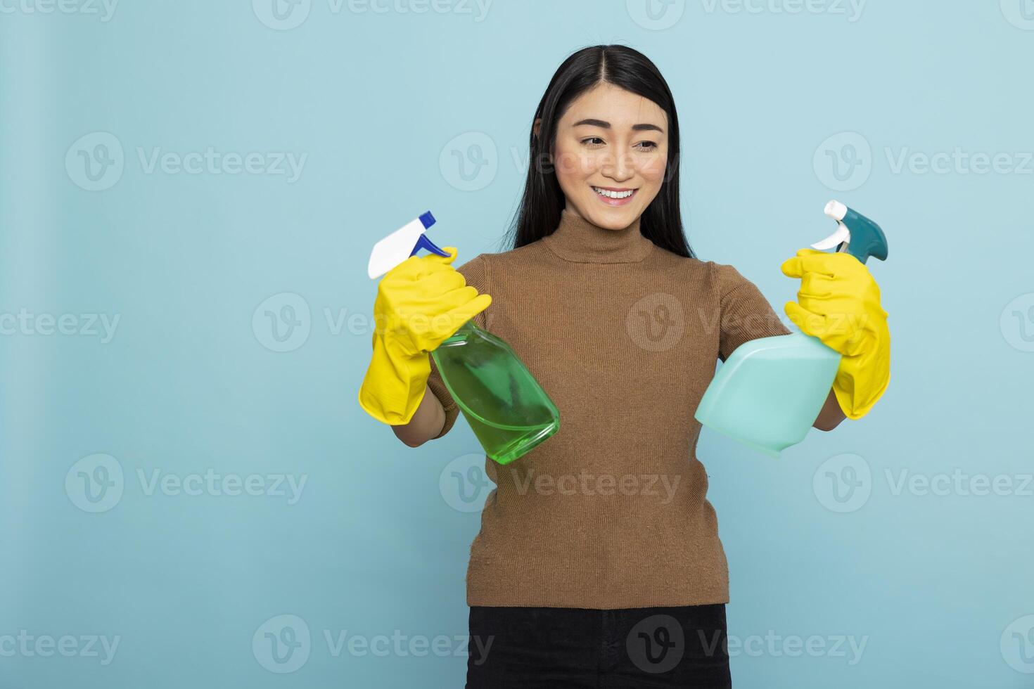 Smiling asian janitor standing against blue background, confidently holding spray bottles and wearing yellow rubber gloves. Happy housekeeper ready to begin morning housecleaning routine. photo
