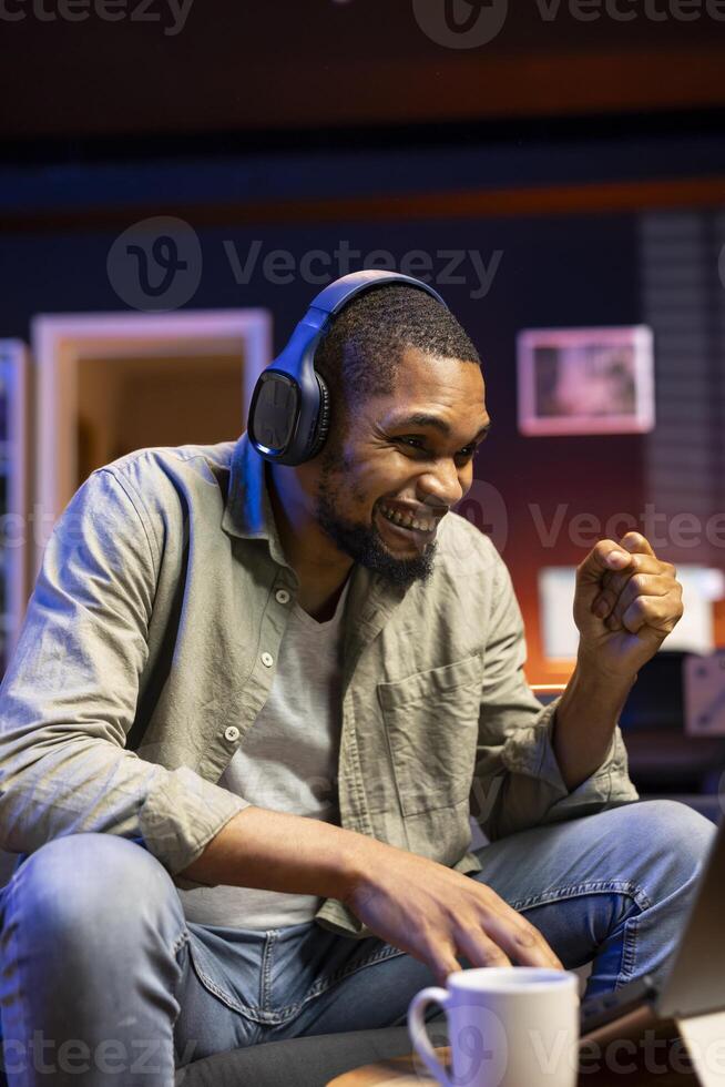 Content cheerful adult enjoying his work progress from his living room, celebrating career achievement and listening to music while solving tasks. African american guy multitasking. photo