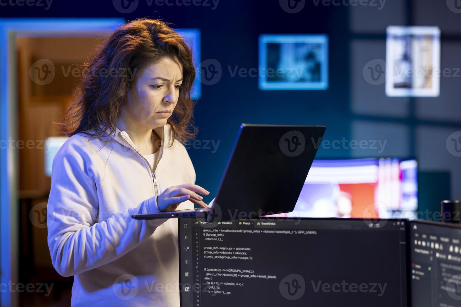 IT system administrator doing software debugging using laptop, checking glitches in lines of code displayed on desktop PC monitors. Woman standing in office at home fixing coding issues using notebook photo