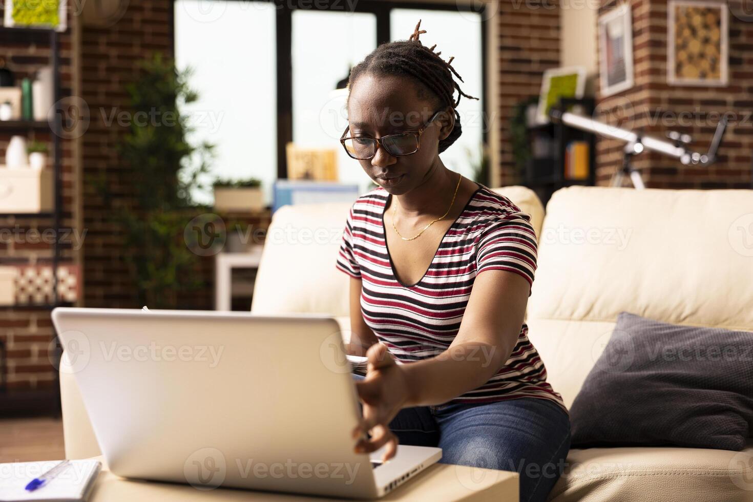 Focused woman works from home, reviewing company growth analytics on laptop. African American female entrepreneur touching device screen and reading online data with full concentration. photo