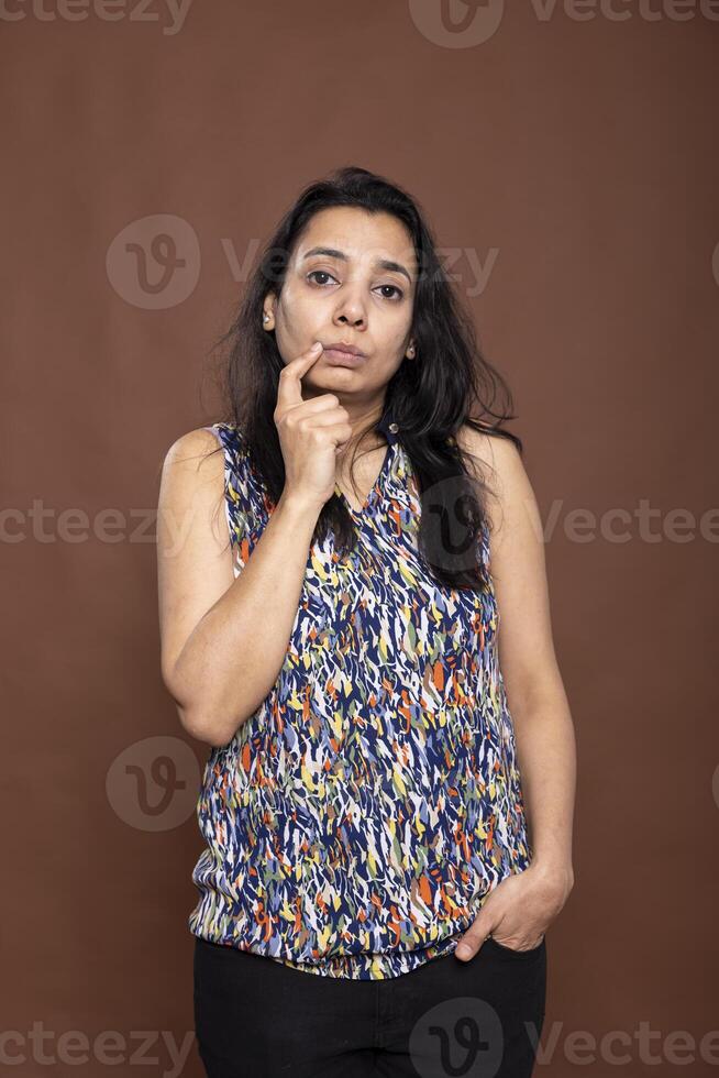 Contemplative woman standing with thoughtful expression, touching her chin and pondering an answer. Female in casual outfit appearing curious and doubtful, isolated in brown studio background. photo