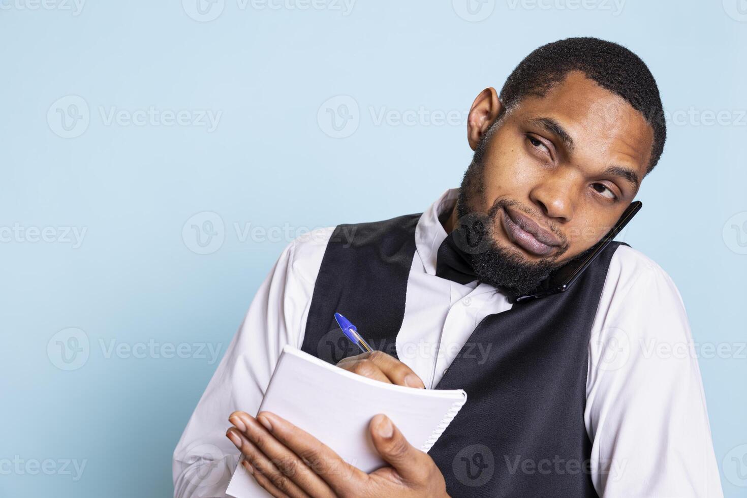 Front desk staff writing down a list of names for room reservations, booking via smartphone call. Receptionist hotel porter takes notes for the requested amenities in studio. photo