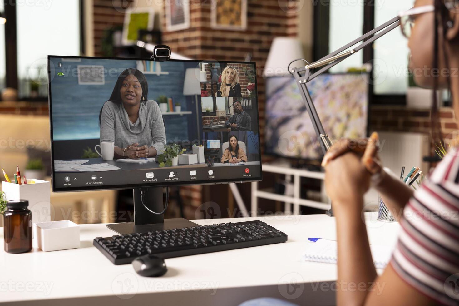 Female entrepreneur works from home, engaging in virtual discussion with diverse business partners. African American woman sits at desk in brick wall room, participating in online training on computer photo