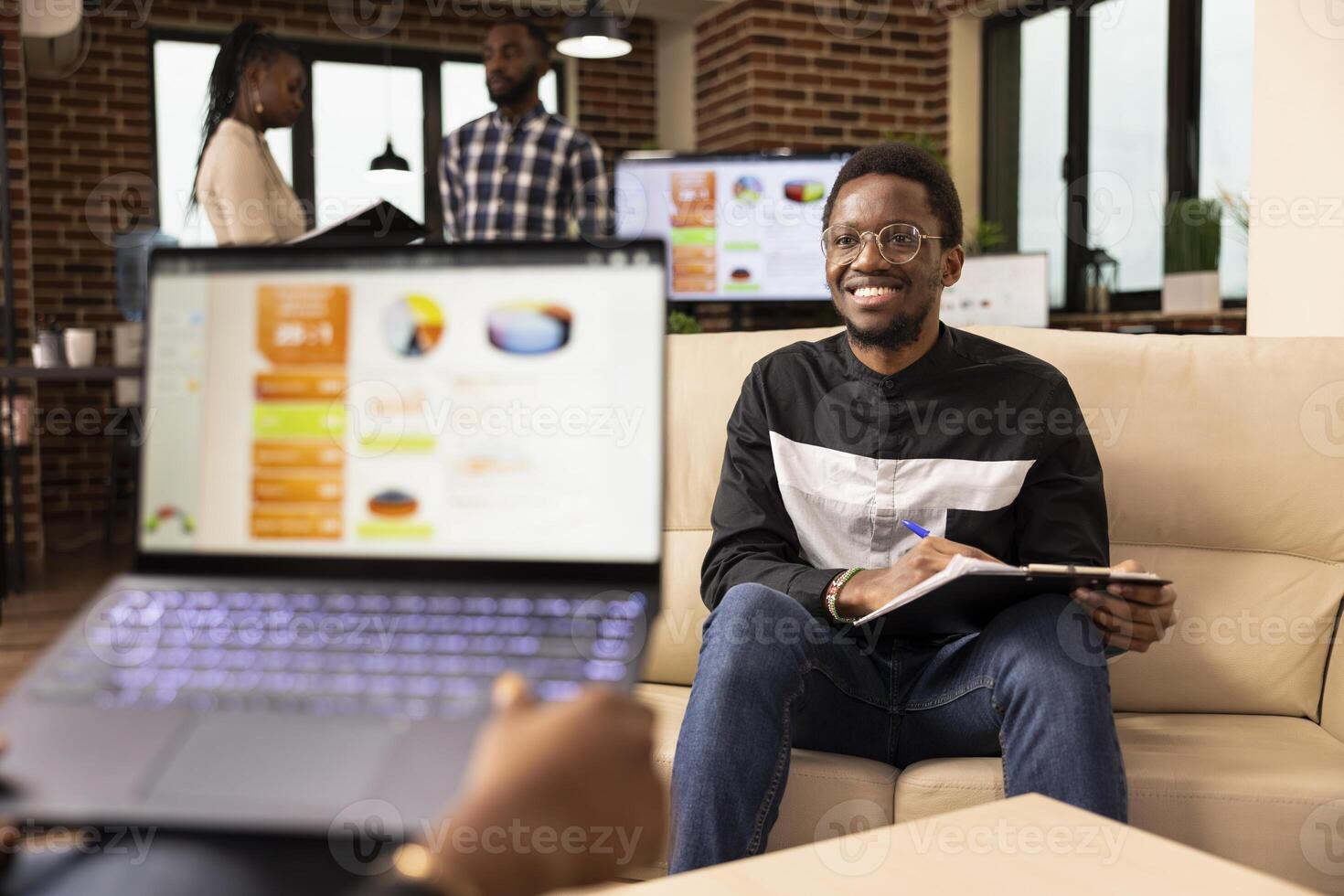 Two business professionals meet in office to share ideas and collaborate on project task. African american sales manager with laptop presents commercial offer to startup company client seated on sofa. photo
