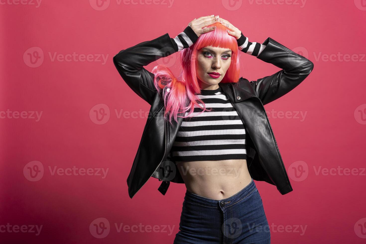 Trendy model with pink hair and bold jacket embraces confident rocker style against isolated background. Pretty woman exudes funky attitude and stands gracefully, expressing punk vibe and chic flair. photo