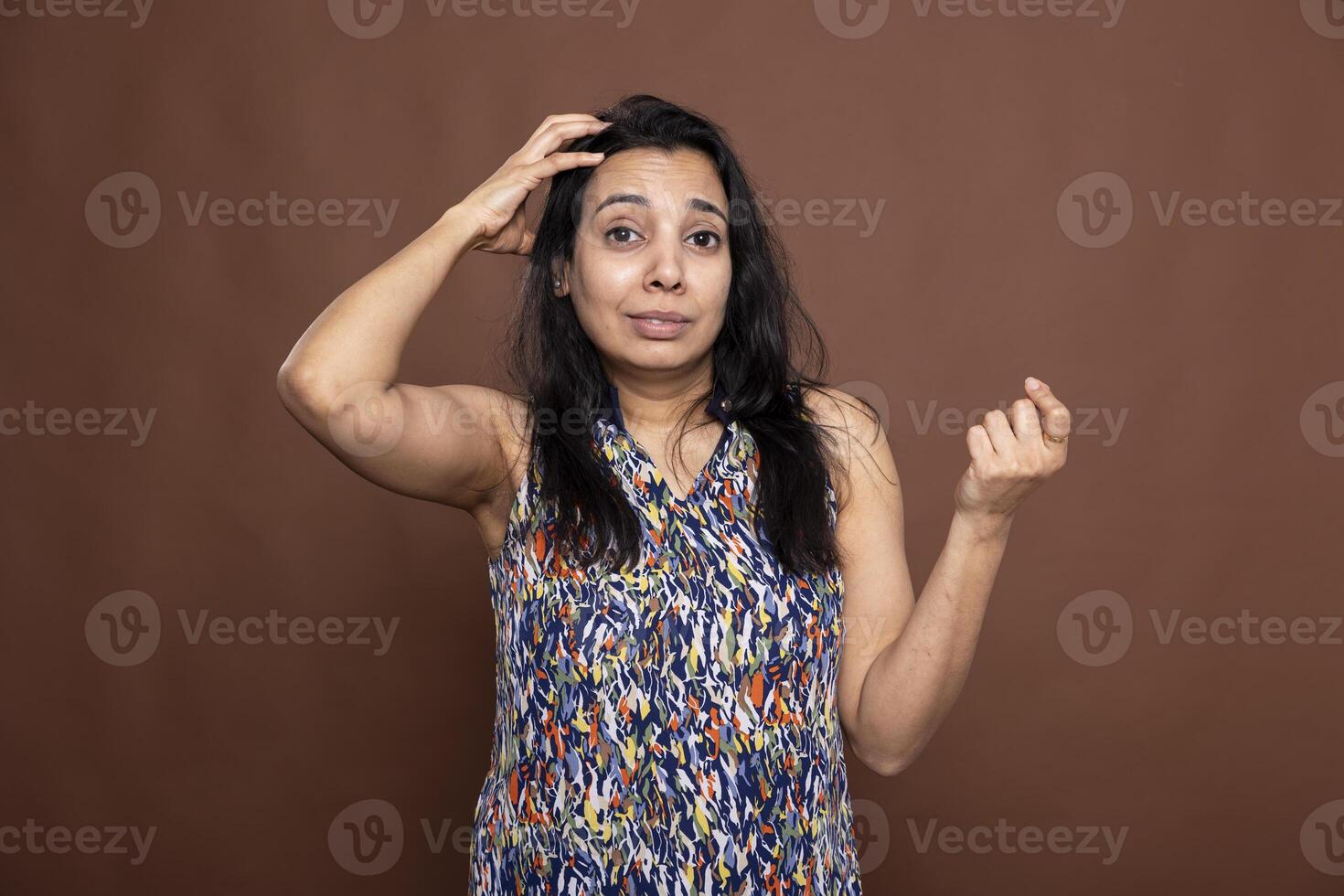 Puzzled adult woman scratching her head and looking at camera with troubled expression in brown background studio. Pretty indian woman appearing confused and indecisive, unsure about something. photo