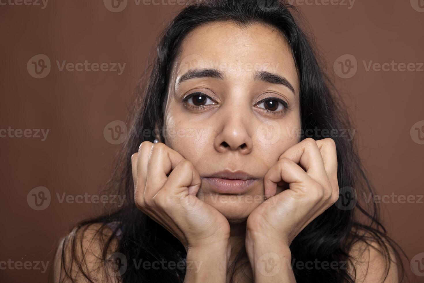 Closeup of sad indian woman looking directly at camera with troubled, distressed expression. Anxious female model resting head on her hands, feeling emotional after receiving upsetting news photo