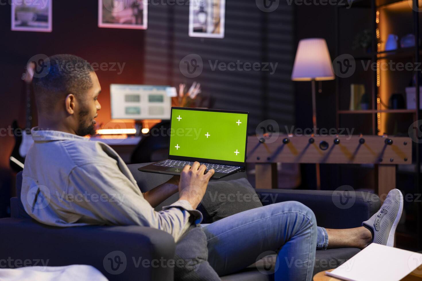 African american young guy using device with green screen from his sofa, enjoys the comfort of his modern cozy home. Casual relaxed person working in the living room, multitasking. photo