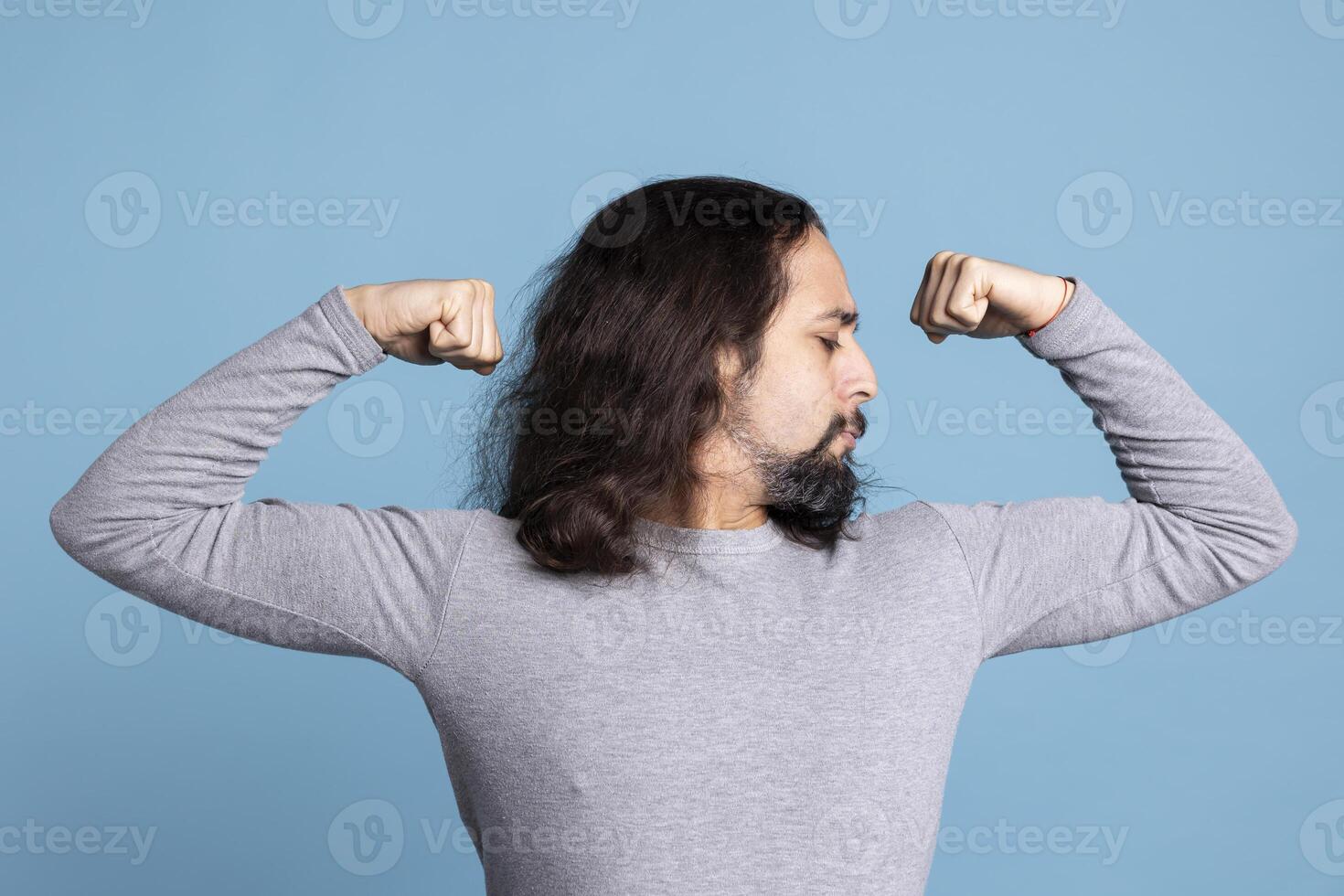Athletic young guy flexing his arms muscles against blue background, showing off his toned fit physique in front of the camera. Fitness motivation and determination from a strong man. photo
