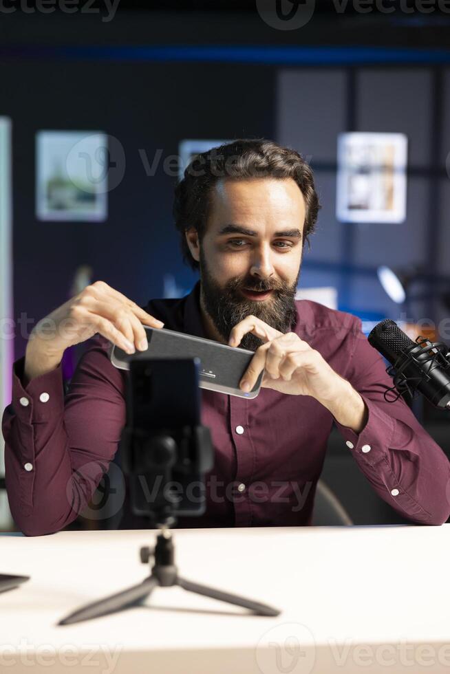 Cheerful online star doing technology review of wireless portable speaker, filming with phone on tripod. Happy influencer showcasing music playing device in apartment studio photo