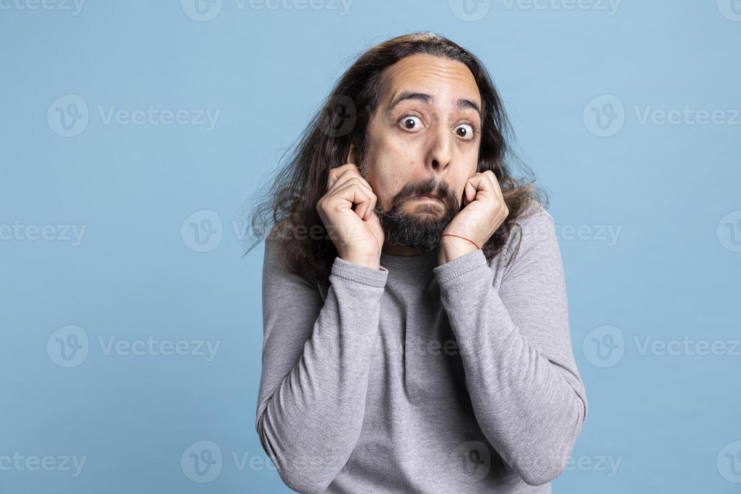Joyful bearded man with long curly hair strikes a silly pose showing a carefree attitude. Playful behavior and comical face highlight his humorous personality in this studio against blue background. photo