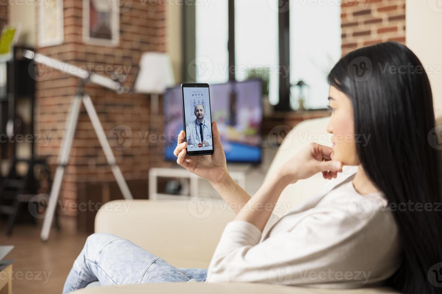 Asian woman sits on couch, using cellphone for remote healthcare session with caucasian physician. Female entrepreneur attending virtual medical consultation from home, speaking with male doctor. photo