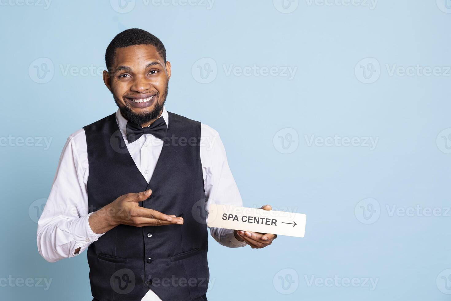 Hotel employee in tuxedo pointing people to the spa center using a wall sign, holding the indicator to guide guests to resort facilities. Offering great services at the five star lodging. photo