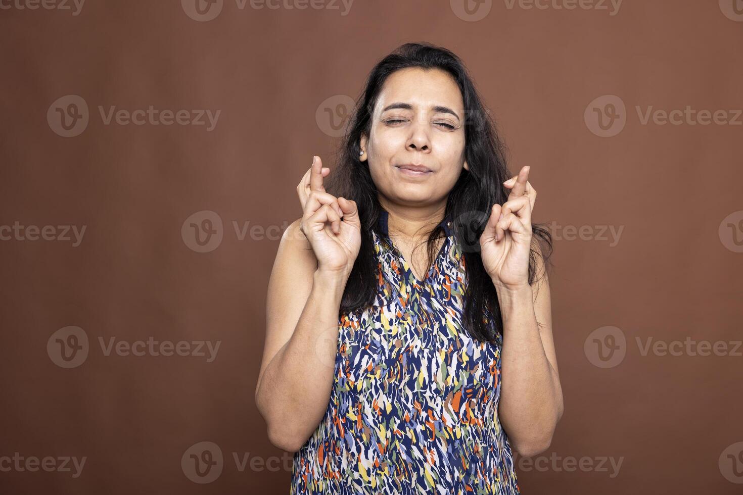 Indian woman stands with eyes closed and fingers crossed, quietly hoping for favorable result. Female individual with pleading expression, silently wishing for good news or miracle. photo