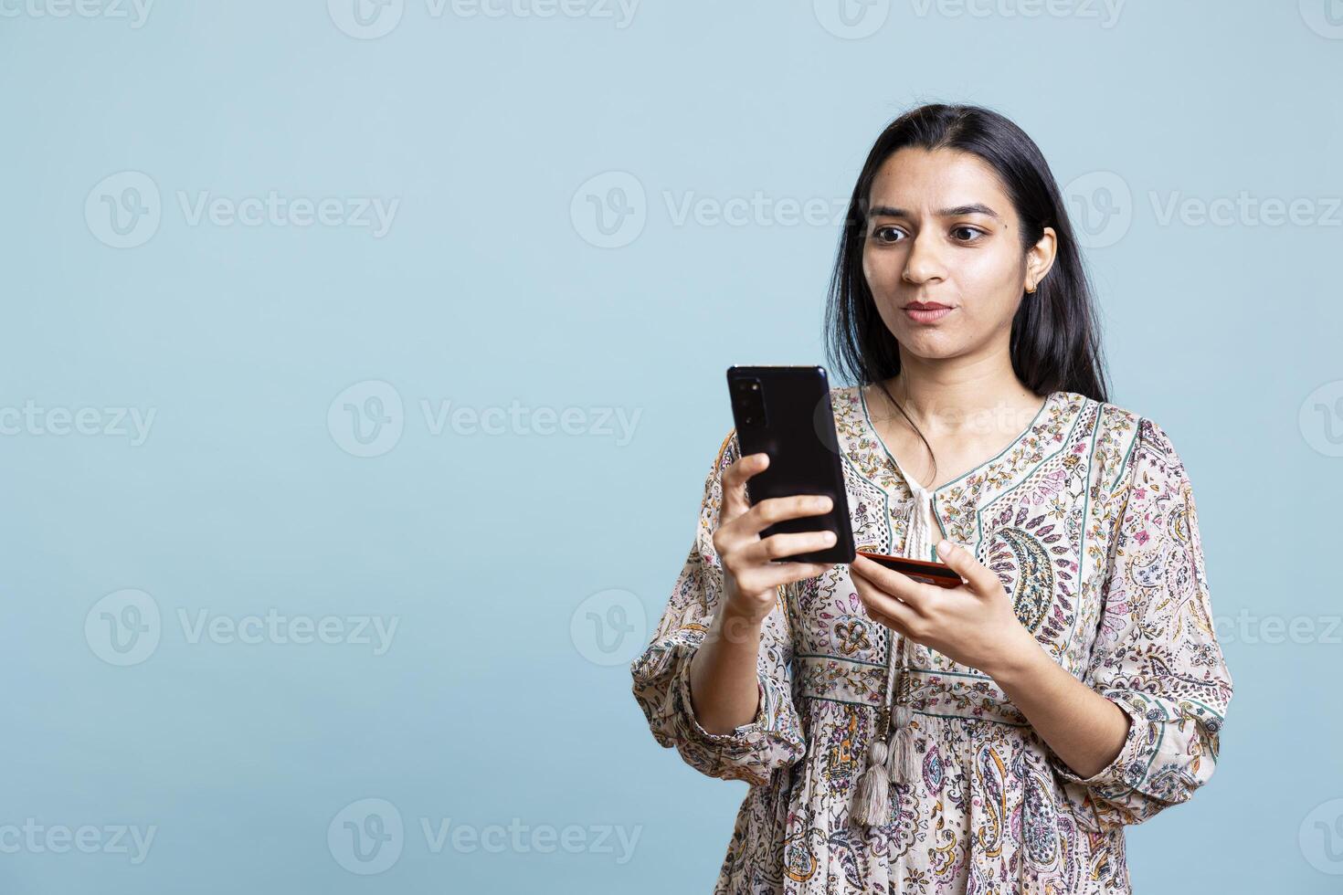 Upbeat person adding banking credentials on her mobile phone, confirming payment and money transfer on a website. Young indian adult buying online using card against studio background. photo