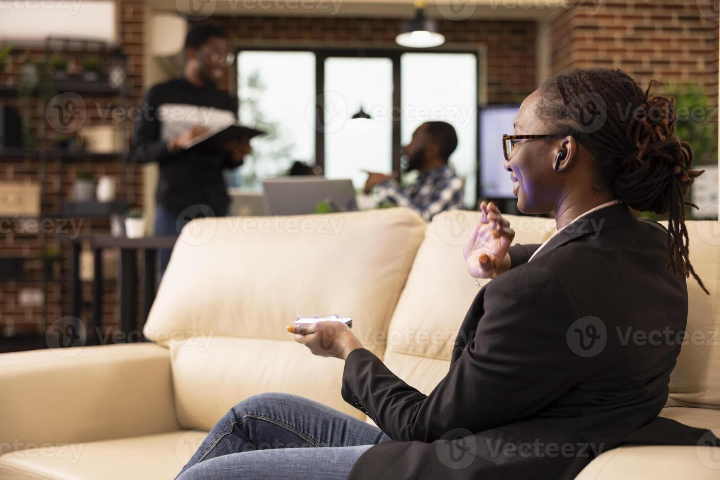 Black female entrepreneur using futuristic device to communicate with holographic interface and present data in tech forward workspace. African american team in background symbolizes startup synergy. photo