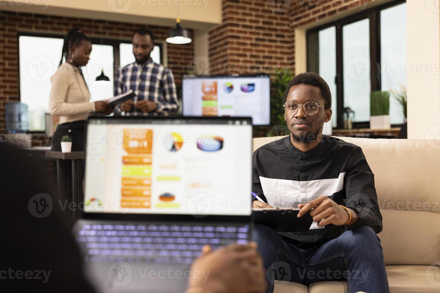 African american manager with laptop explains business plans and solutions to young male employee. Black businessman sitting on sofa with clipboard, attentively listens to coworker during meeting. photo