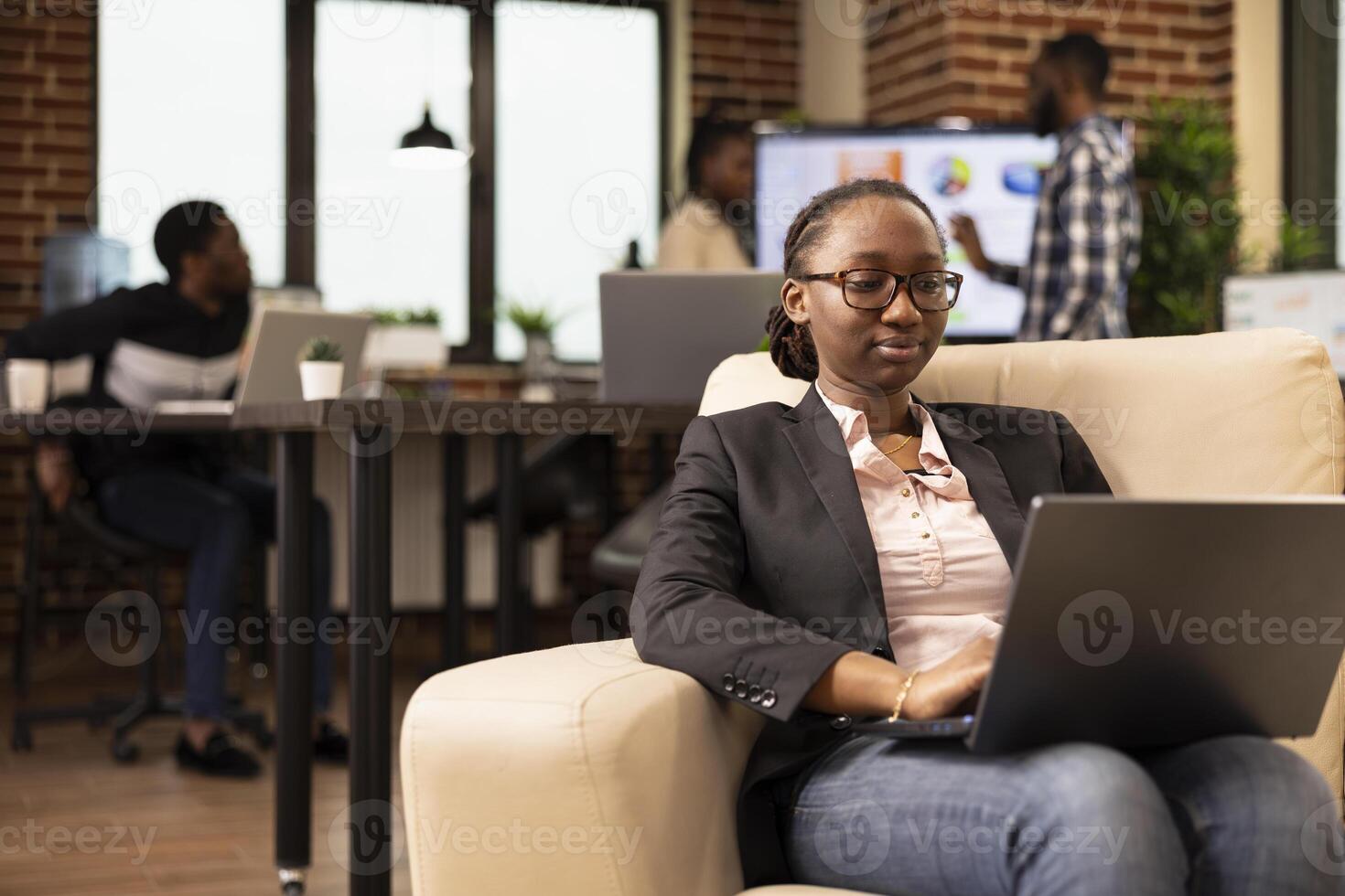 Black female company advisor using laptop, seated on couch, analyzing marketing strategy and compiling reports. African american woman manager comfortably working on business project in startup office photo