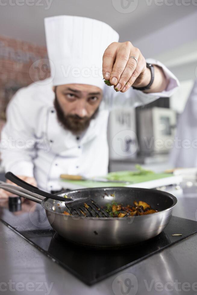Professional chef in a modern kitchen carefully sprinkles fresh herbs onto a dish, enhancing the flavors with parsley, rosemary and basil. Modern workspace filled with fresh produce and spices. photo