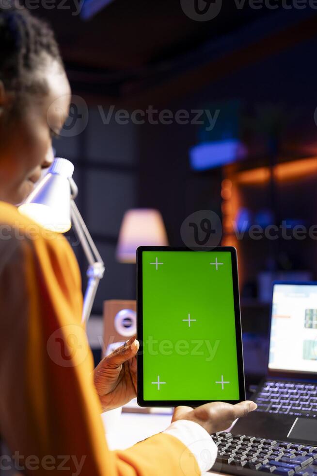 Woman sitting on chair at home office desk in neon illuminated apartment holding green screen tablet while working. Person looking at business graphs on laptop and at mockup device photo