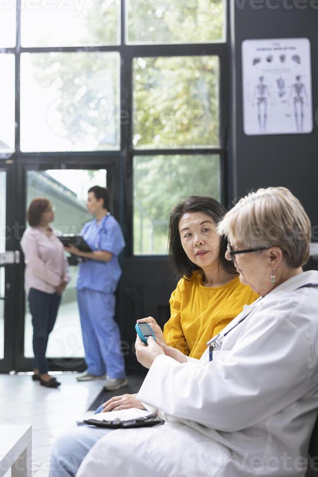 Older physician using a glucometer for blood sugar monitoring, doing a routine checkup for asian patient suffering from diabetes. Measuring glucose and sugar levels. Device training. photo