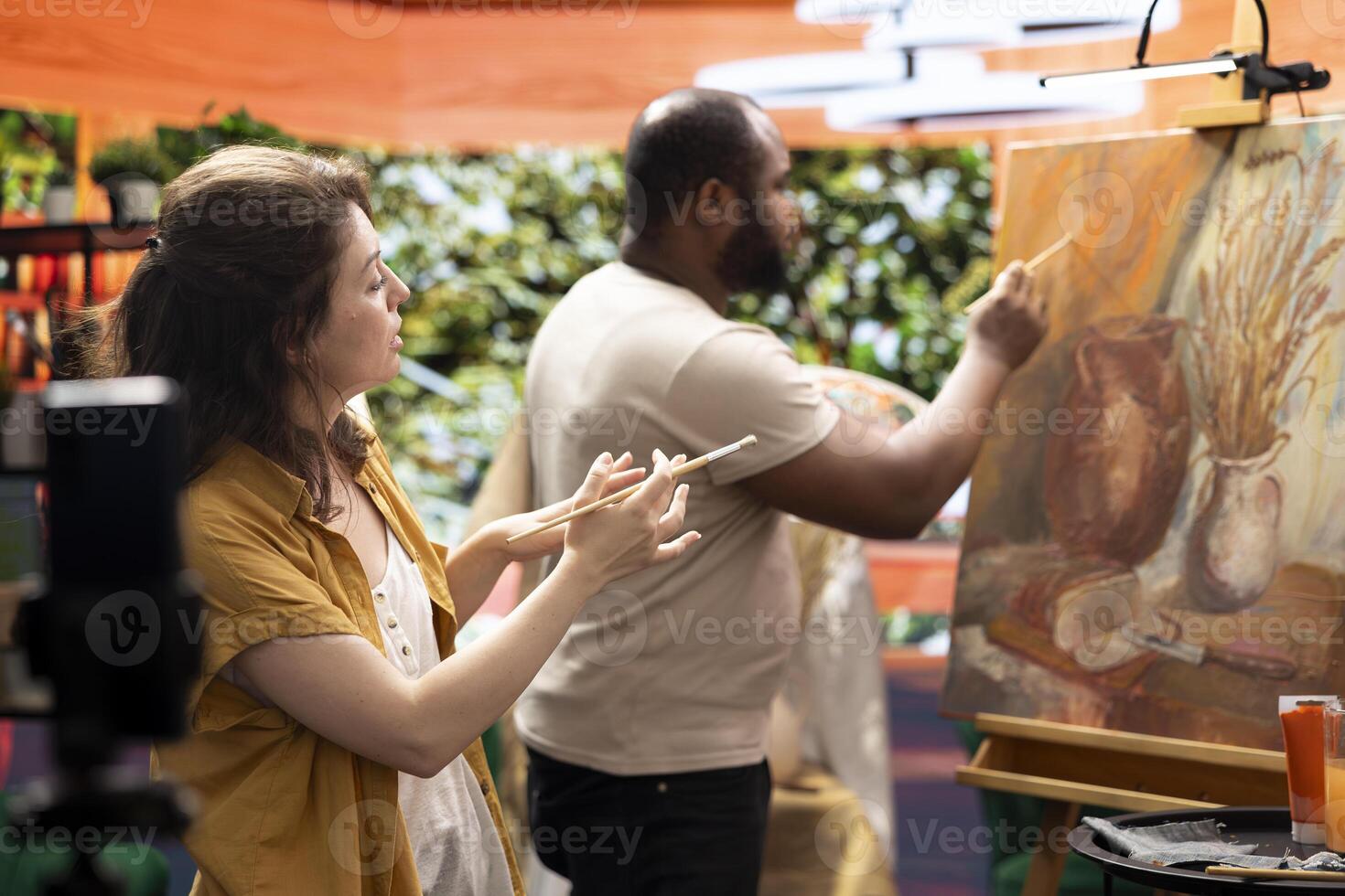 Woman painter filming her colleague during his creative process and explaining the painting technique for online tutorial. Diverse artists creating virtual classroom to teach art. Digital education. photo