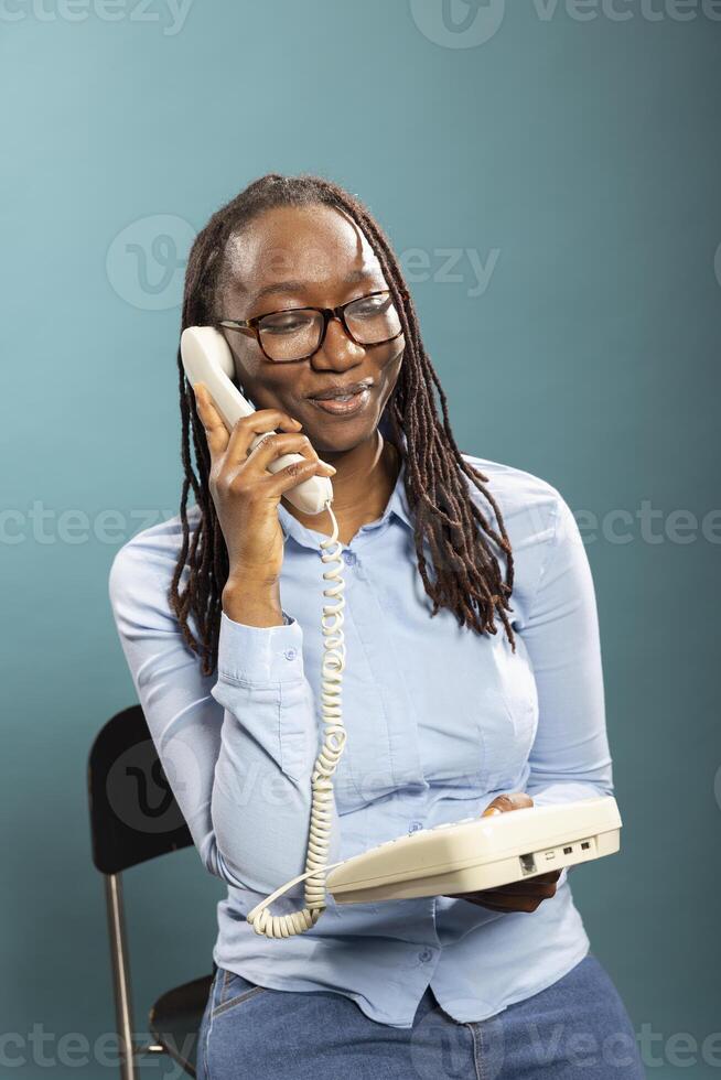 Portrait of smiling black woman using telephone, talking with family and friends in modern studio. African american female model holding landline receiver to her ear, having a phone call. photo