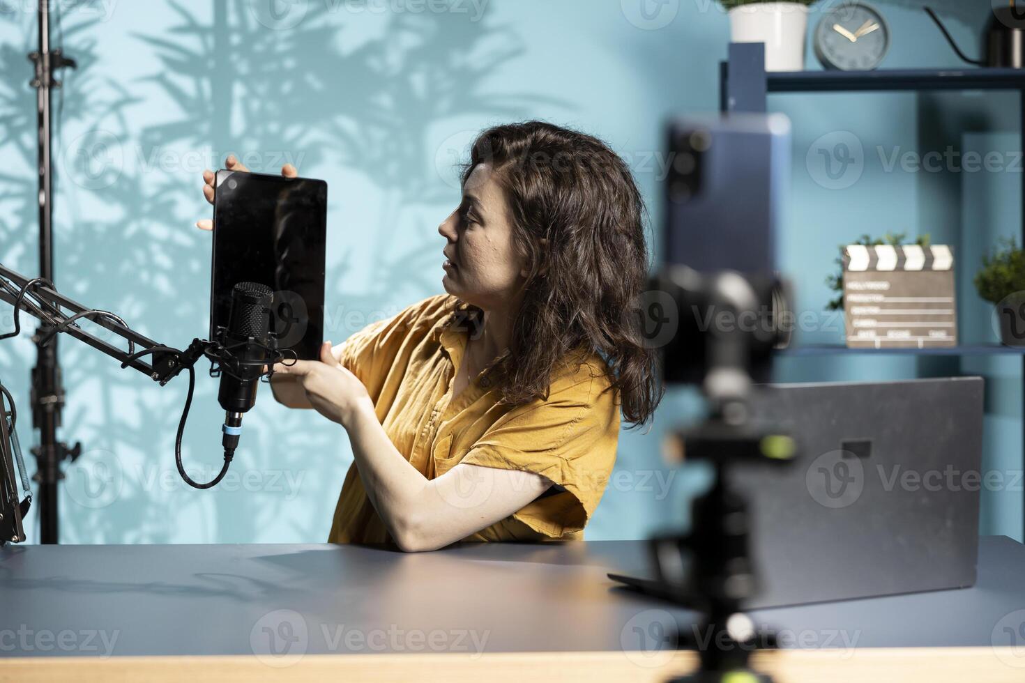 Tech specialist presenting tablet in entry level low budget price range to interested viewers, filming with smartphone on tripod. Woman reviewing digital device, recording with mobile phone photo