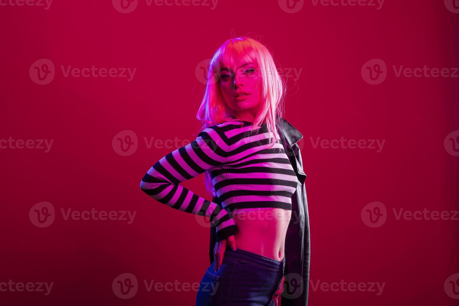 Confident female model strikes a pose with hand on waist under vibrant red pink studio lights. Gorgeous woman wearing striped top and dark leather jacket, enhancing the rebellious fashion concept. photo