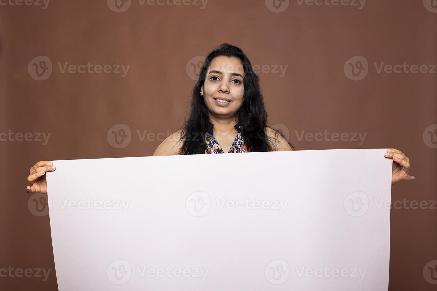 Indian woman stands holding blank white banner, ready for advertisement use against brown background studio. Female individual posing confidently, grasping empty placard, perfect for promotional ads. photo