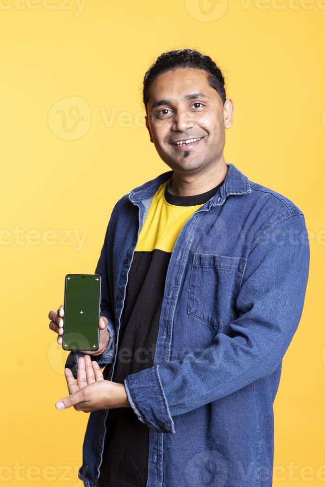 Portrait of indian person doing ad marketing using isolated chroma key on gadget, guy with man bun standing against studio background. Smiling man pointing at empty green screen. photo
