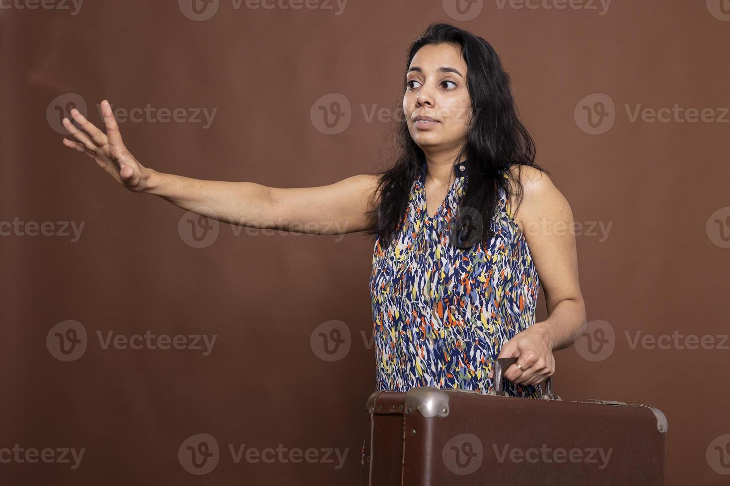 Beautiful indian lady with one arm outstretched and other carrying suitcase, portraying lateness, rushing to catch flight. Portrait of female tourist with luggage, expressing sense of urgency. photo