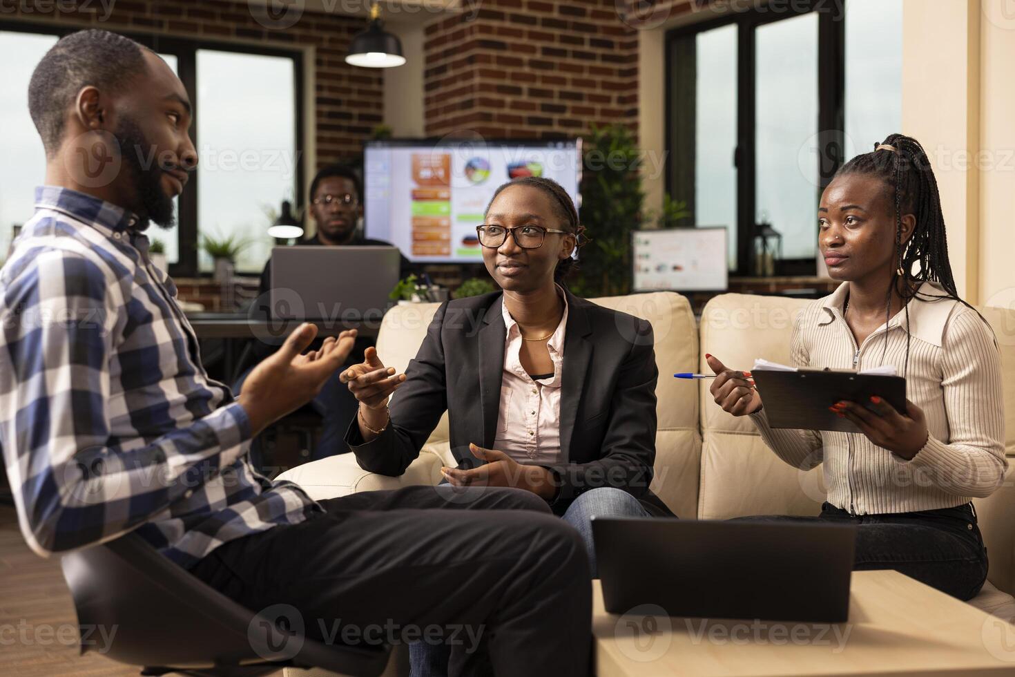 African American coworkers sitting in office with laptop and clipboard, reviewing paperwork and discussing financial project report. Busy startup team collaborates efficiently in a modern workspace. photo