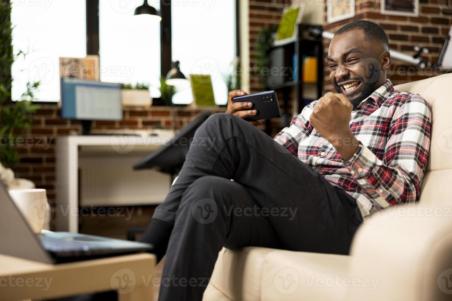 Cheerful male entrepreneur on couch, celebrating with fist raised after winning mobile game. Smiling businessman resting from remote work, holds cellphone, feeling excited from online gaming success. photo