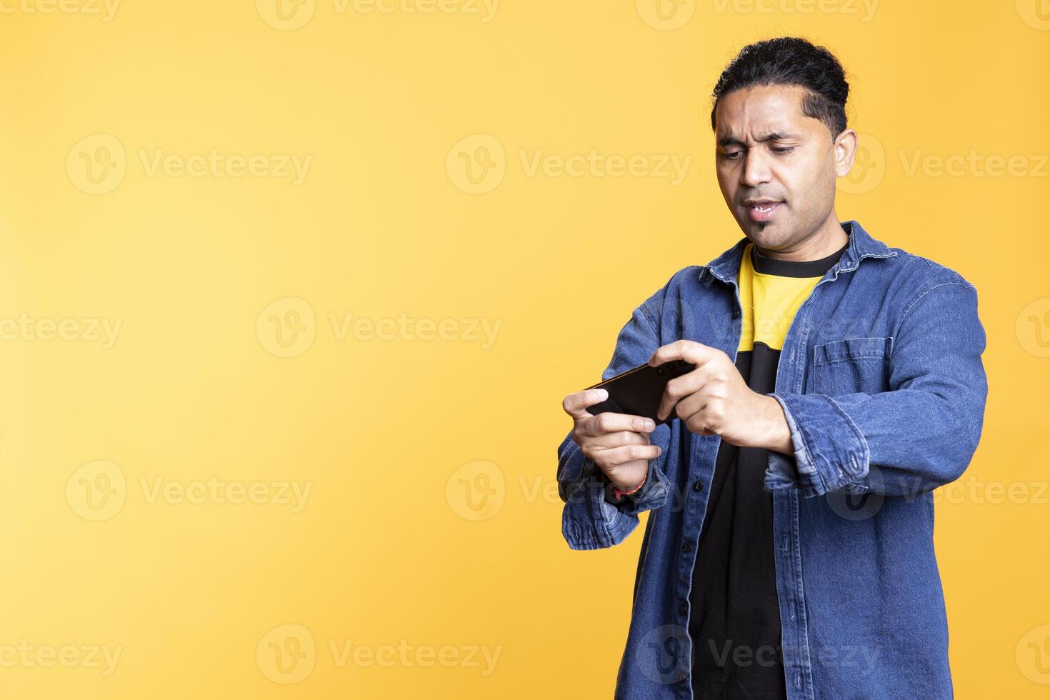 Relaxed Indian guy being entertained by games on mobile phone, enjoying leisure time in studio. Young player having fun playing games and defeating enemies, man bun expression. photo