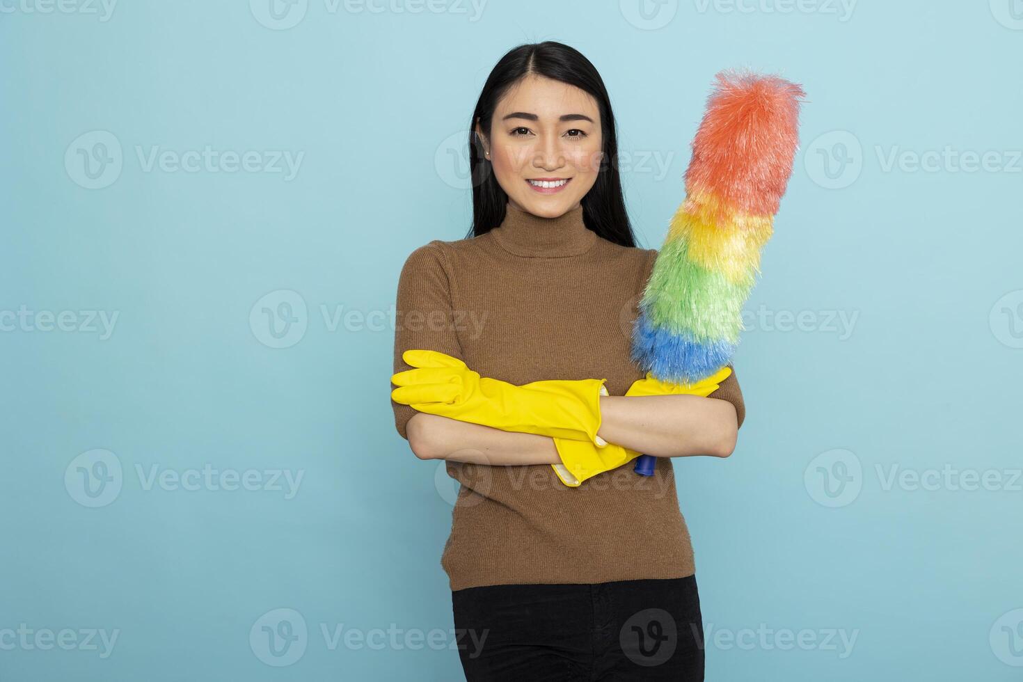 Happy young asian housekeeper wearing yellow rubber gloves, smiling confidently with feather duster in hand. Female cleaner standing with arms crossed, ready for household cleaning tasks. photo