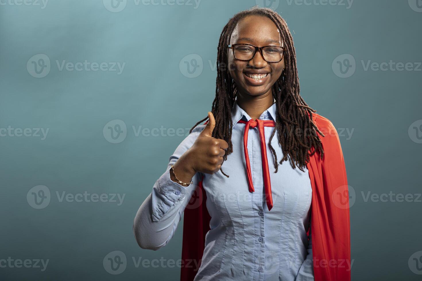 Cheerful black heroine giving a thumbs up, portraying positivity and agreement. African american woman in red cape showing approval gesture and looking at camera with bright smile. photo