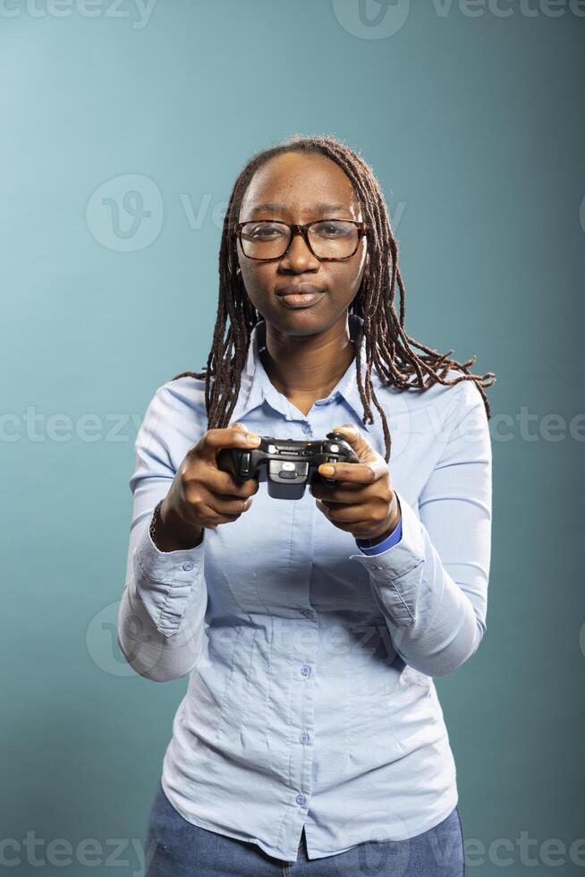 Black woman with glasses holding gaming controller and having fun playing games in modern studio. African american female model in blue shirt fully engaged in a serious gaming session. photo