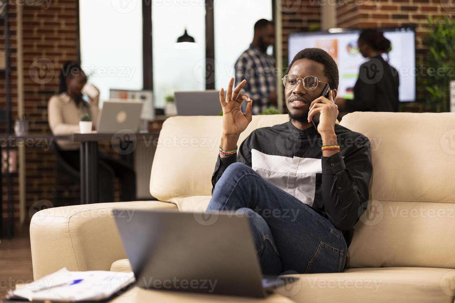 Black male professional in smart casual clothing, sitting on couch and having an important phone conversation with potential client. African american businessman using smartphone in modern workspace. photo