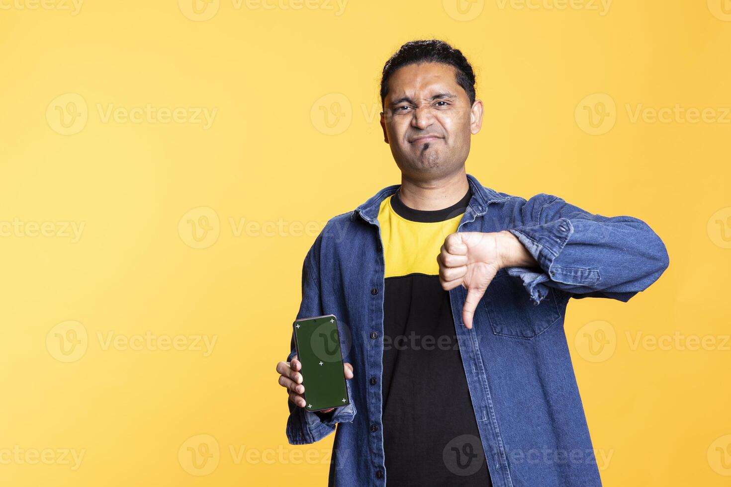 Casual person feels displeased with a thumbs down next to the copy space, holding mobile phone with isolated screen. Indian young adult in the studio, hand gesture with chroma key. photo