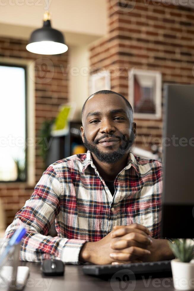 Smiling black remote worker sitting at desk with desktop pc in cozy brick wall living space. Self employed male professional prepared for virtual meetings and business tasks from home. photo