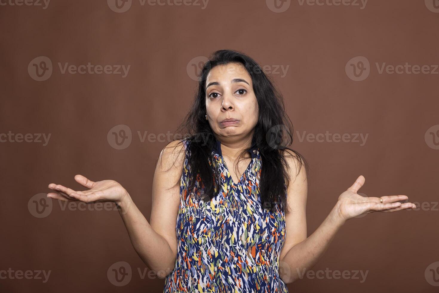 Portrait of indian woman shrugging shoulders with puzzled expression, standing against brown background. Beautiful female model holding hands out, clearly unsure and making I dont know gesture. photo