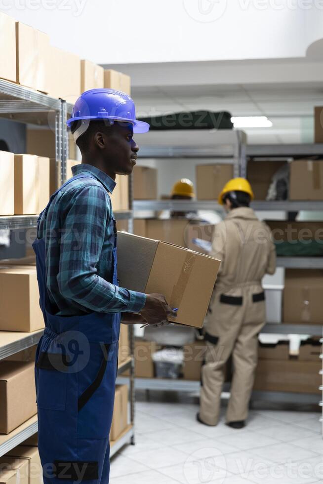 African american worker in protective gear checks merchandise for dispatch in a busy warehouse. Crates and boxes arranged for logistics and transportation in operational supply chain hub. photo