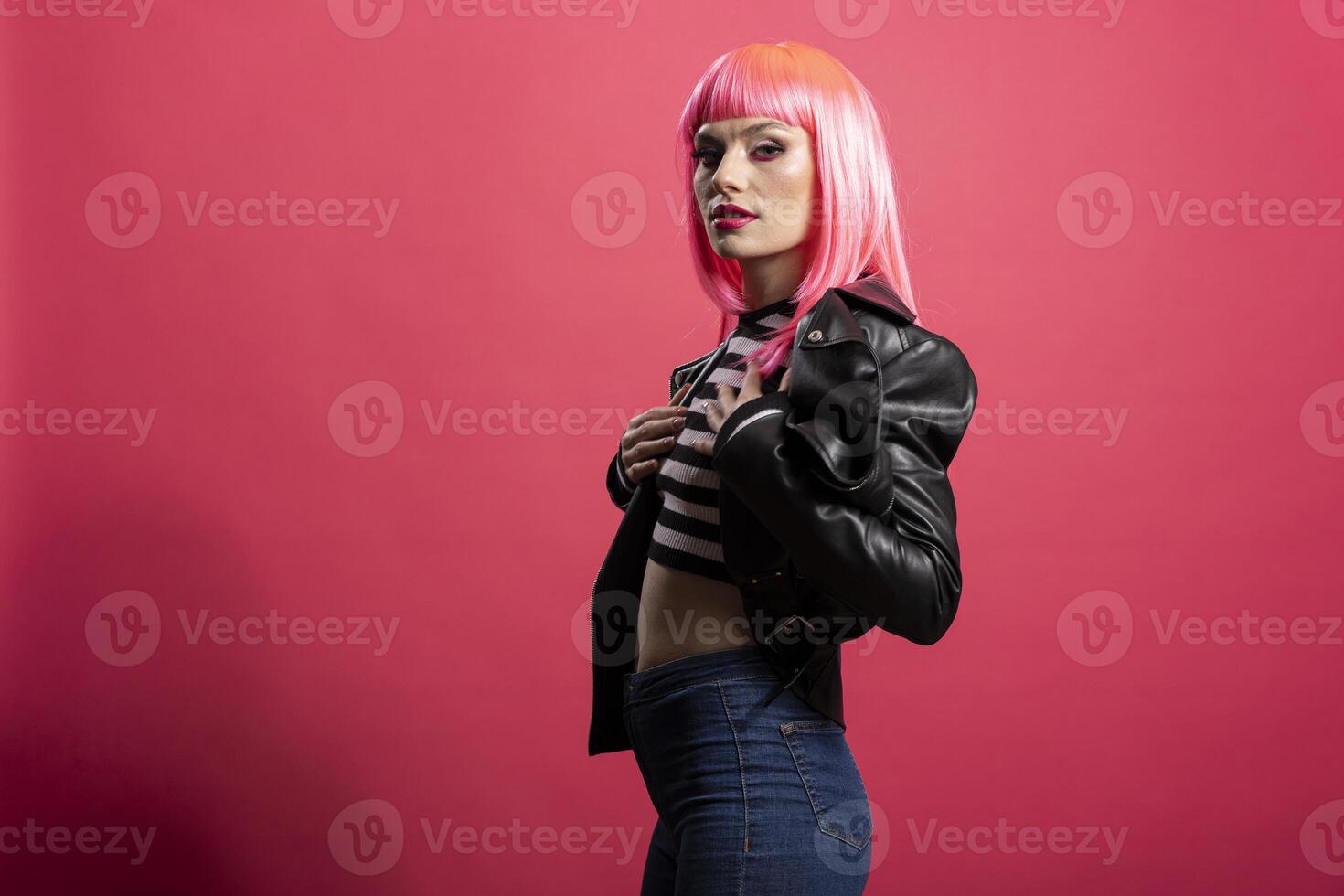 Stylish fashion model wearing striped crop top and leather jacket, glowing with beauty while staring directly at camera with focused gaze. Beautiful carefree woman stands isolated over pink background photo