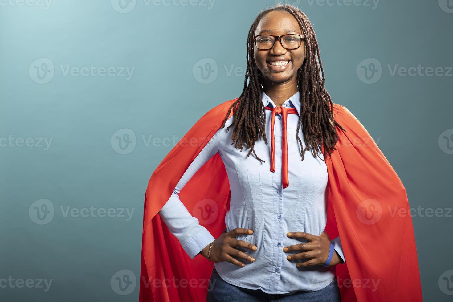 African american woman in red cape and blue shirt, hands on waist, smiling brightly at camera. Black female model stands in a heroic pose, portraying a confident corporate superhero. photo