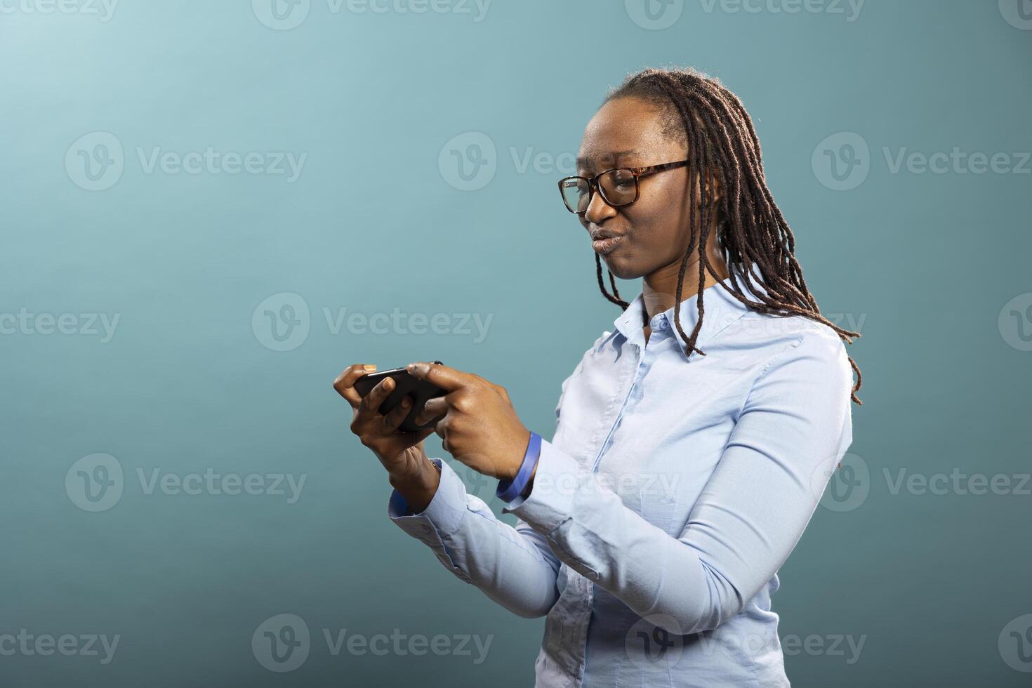 African American woman in blue shirt focuses intently on her smartphone, watching a in studio. Black female individual stands isolated over blue background, using her mobile phone. photo