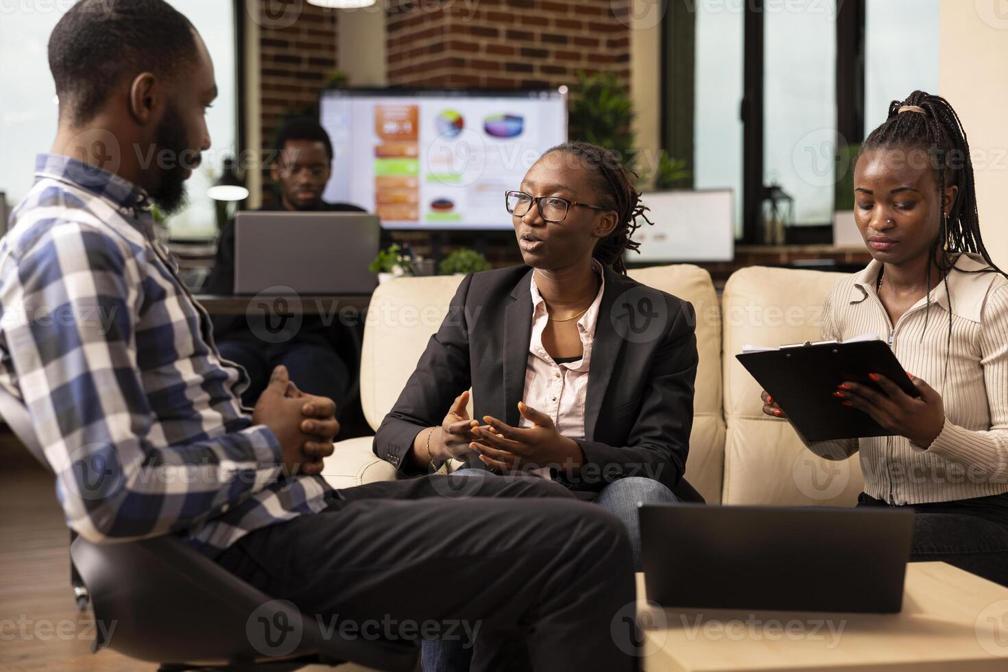 Professional woman with glasses providing business project updates to male colleague during meeting. African american coworkers seated at office lounge area, discussing marketing plans and solutions. photo
